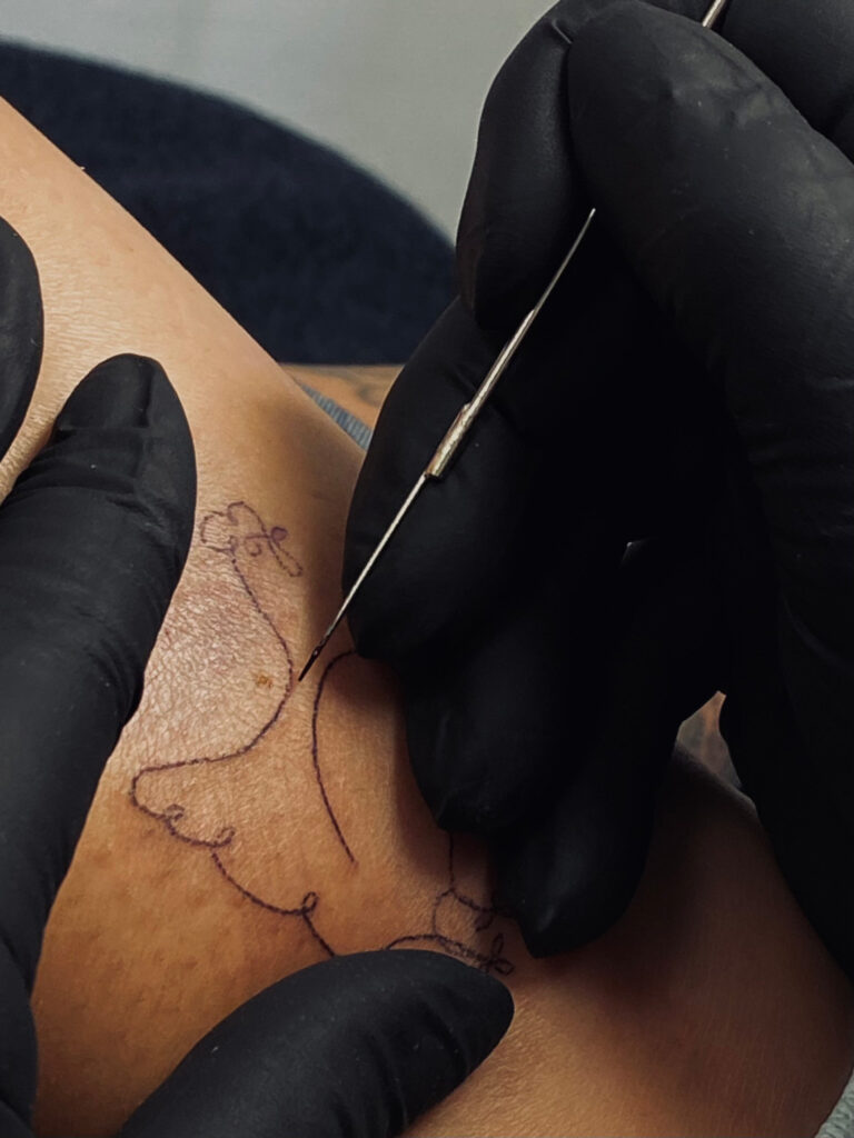 Tattoo wird gestochen auf einem Arm; schwarze Handschuhe; dezentes Linienmuster.